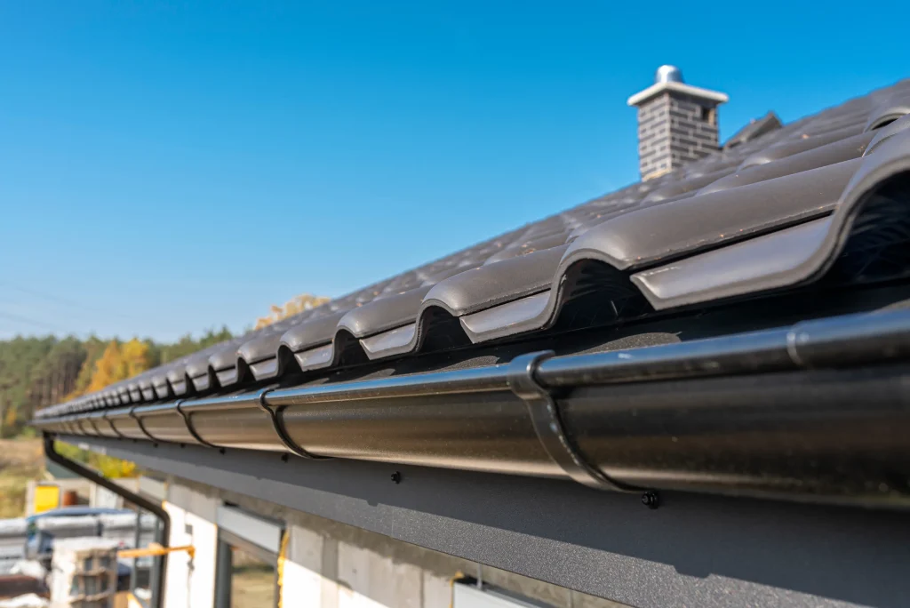 Close-up of a modern dark roof with sleek black gutters against a clear blue sky. A chimney is visible, and trees are in the distant background.