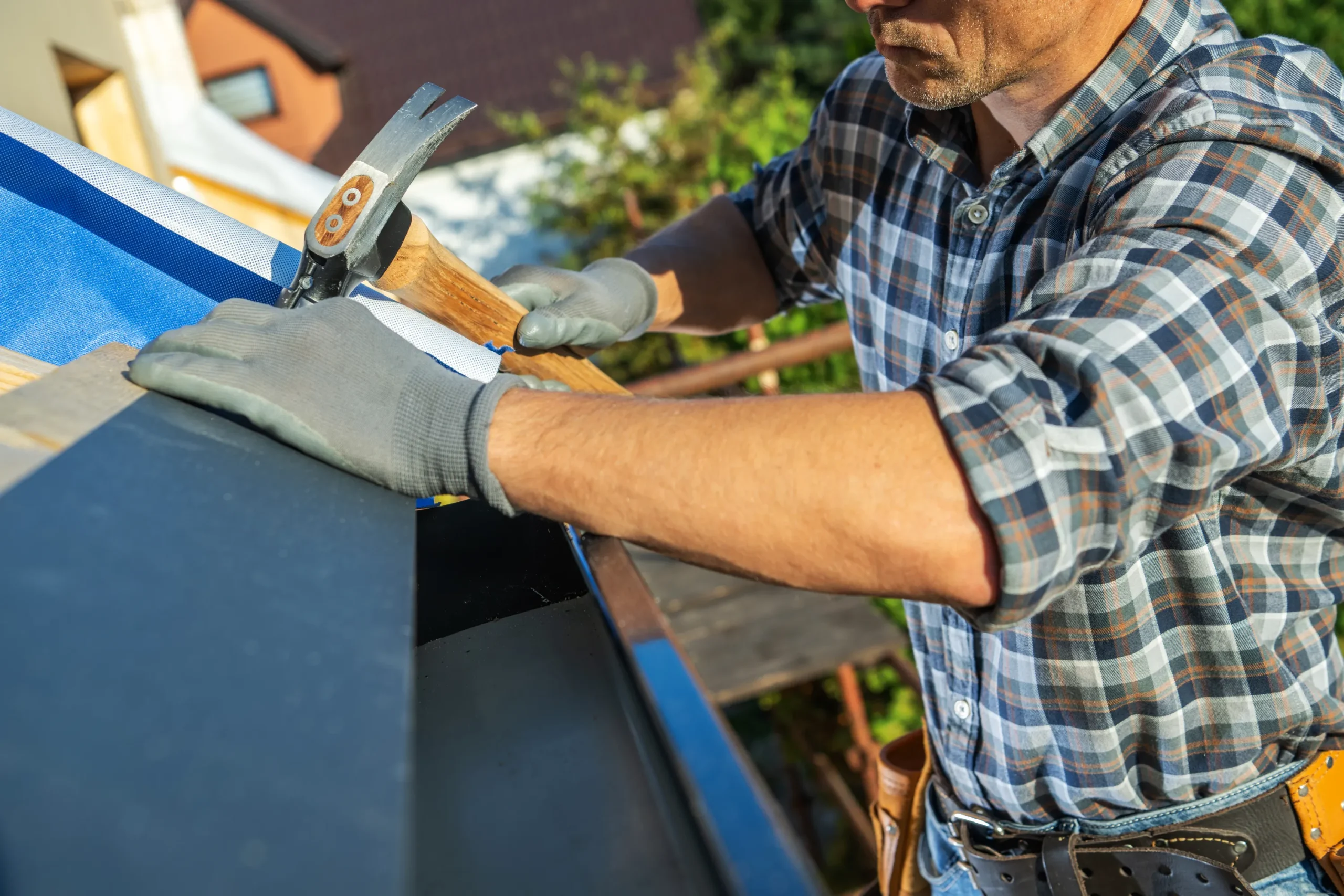 Man in plaid shirt and gloves hammering on a rooftop, surrounded by greenery. The scene conveys a sense of focused craftsmanship.