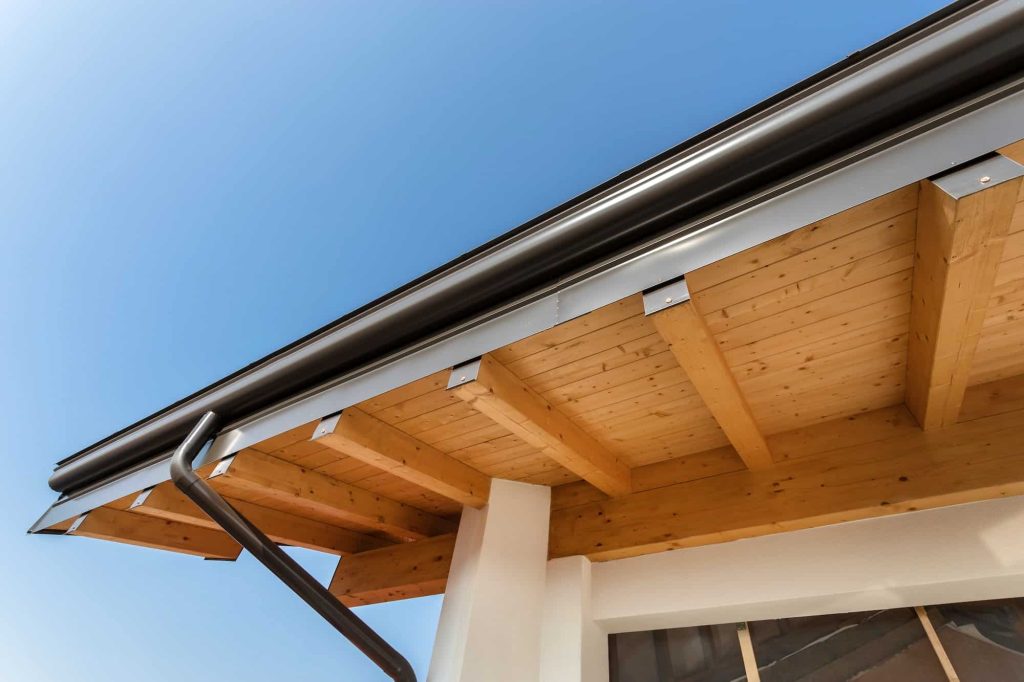 Close-up of a house roof corner with light wood beams and a sleek, black gutter under a clear blue sky, conveying a clean and modern architectural style.