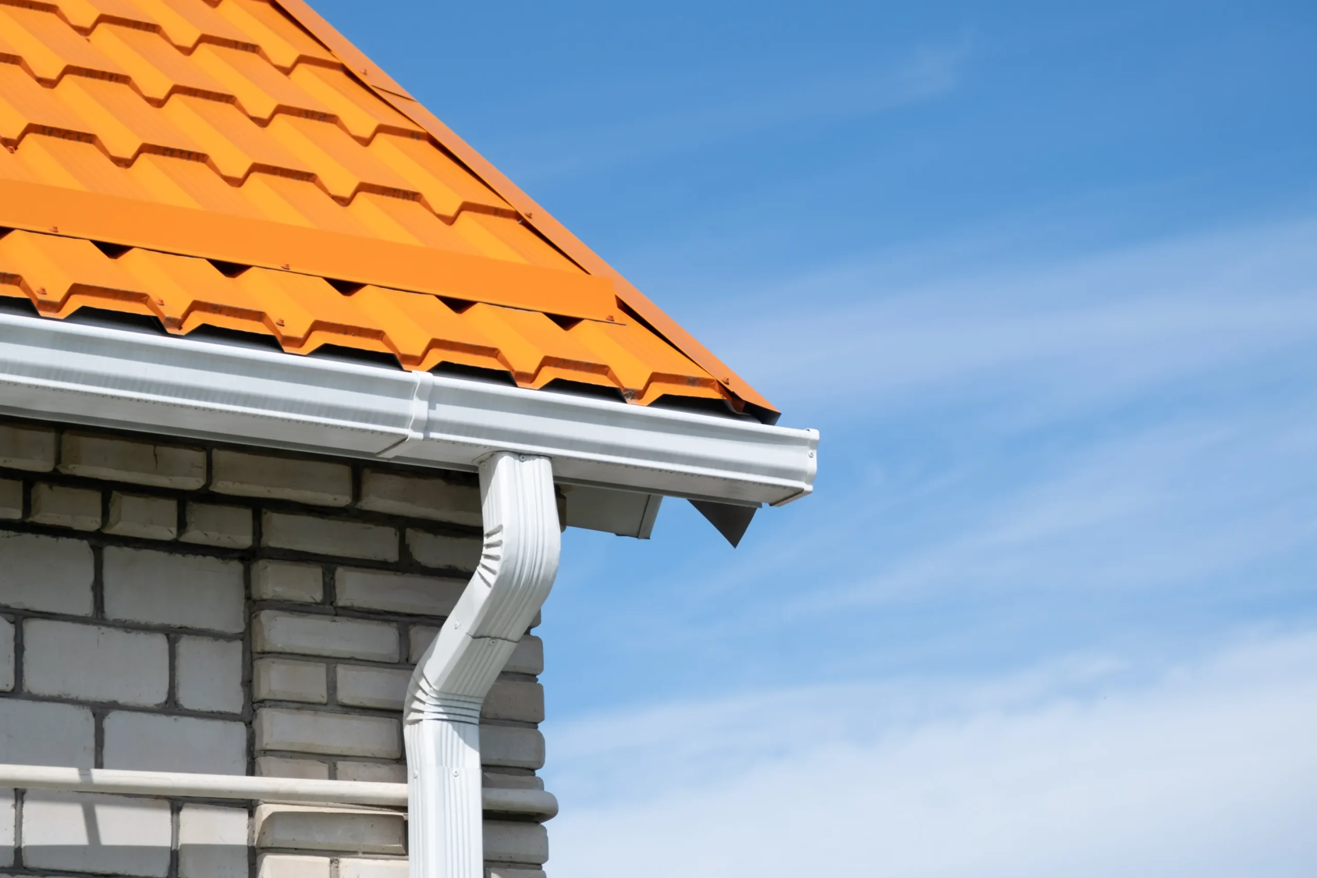 Close-up of a building's corner with orange metal roof tiles, white brick walls, and a silver gutter. The sky is bright blue with wispy clouds.
