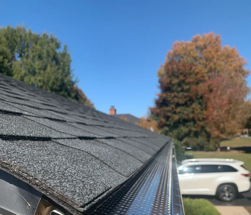 Close-up of a roof edge with dark shingles and a newly installed metal gutter guard. A white SUV is parked on the driveway under a clear blue sky.
